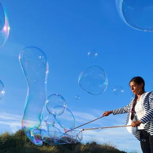 Peut inclure: Une personne souffle de grandes bulles iridescentes contre un ciel bleu clair. Les bulles varient en taille et en forme, certaines étant allongées. La personne porte une veste rayée et tient une baguette à bulles.