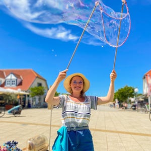 May include: A woman in a straw hat and glasses holds up a large bubble wand, creating a giant bubble against a blue sky. She wears a striped shirt, denim shorts, and a blue scarf. The scene is outdoors on a sunny day.