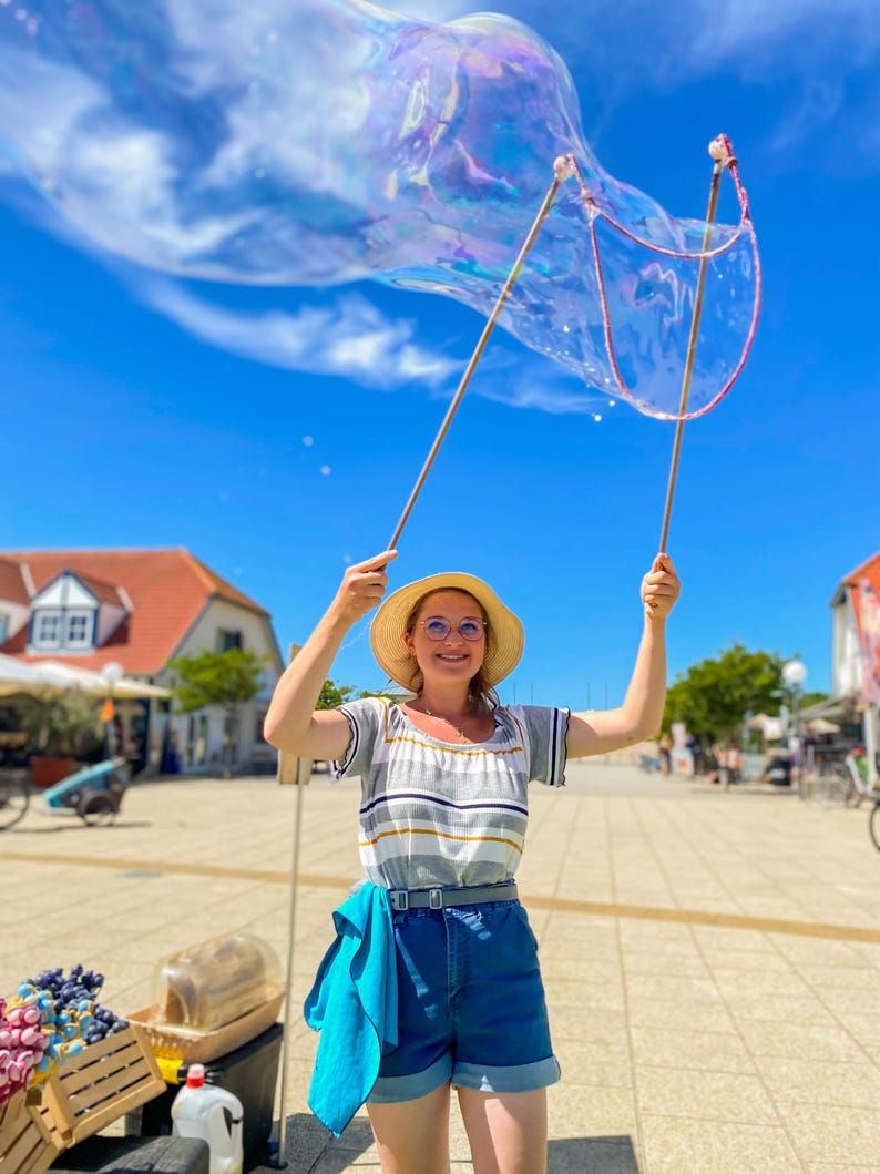 May include: A woman in a straw hat and glasses holds up a large bubble wand, creating a giant bubble against a blue sky. She wears a striped shirt, denim shorts, and a blue scarf. The scene is outdoors on a sunny day.