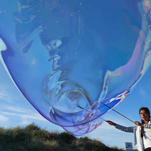 May include: A giant, iridescent blue bubble is held aloft against a clear blue sky. A person in a striped top and white shirt is seen holding the bubble with sticks. The bubble's surface shimmers with colours, reflecting the sky. The background features a grassy hill.