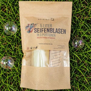 May include: A brown paper bag of bubble solution, with the text "5 Liter Seifenblasen" and "Mische einfach mit Wasser". The bag contains a clear, rectangular block and small packets. Three iridescent bubbles are scattered on the green grass.