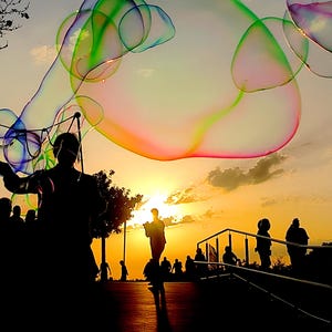 Pode incluir: Grandes bolhas de sabão iridescentes flutuam no ar contra um céu vibrante ao pôr do sol. As bolhas exibem cores rodopiantes de verde, rosa e azul. Silhuetas de pessoas são visíveis numa passarela sob o sol poente no horizonte.