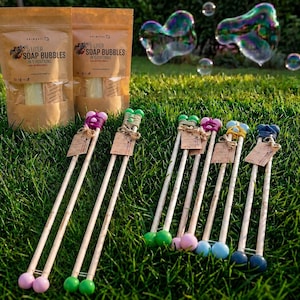 May include: Two brown paper pouches labelled 5 Litre Soap Bubbles in 5 Portions and several bubble wands with wooden handles, colourful yarn accents, and small wooden beads, arranged on green grass with large iridescent soap bubbles in the background.