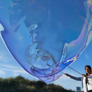 May include: A large, iridescent blue bubble floats in the sky, held by a person with black and white striped sleeves. The bubble reflects the blue sky and has a shimmering, colorful surface. The person is holding two sticks to control the bubble.