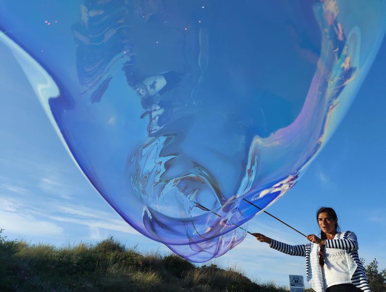 May include: A large, iridescent blue bubble floats in the sky, held by a person with black and white striped sleeves. The bubble reflects the blue sky and has a shimmering, colorful surface. The person is holding two sticks to control the bubble.