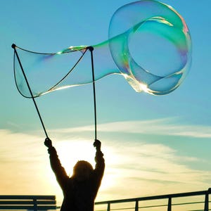 May include: A large, iridescent bubble floats in the sky, held aloft by a person using a bubble wand. The bubble is a vibrant display of colors against a blue sky, with the sun setting in the background. The scene evokes a sense of joy and play.