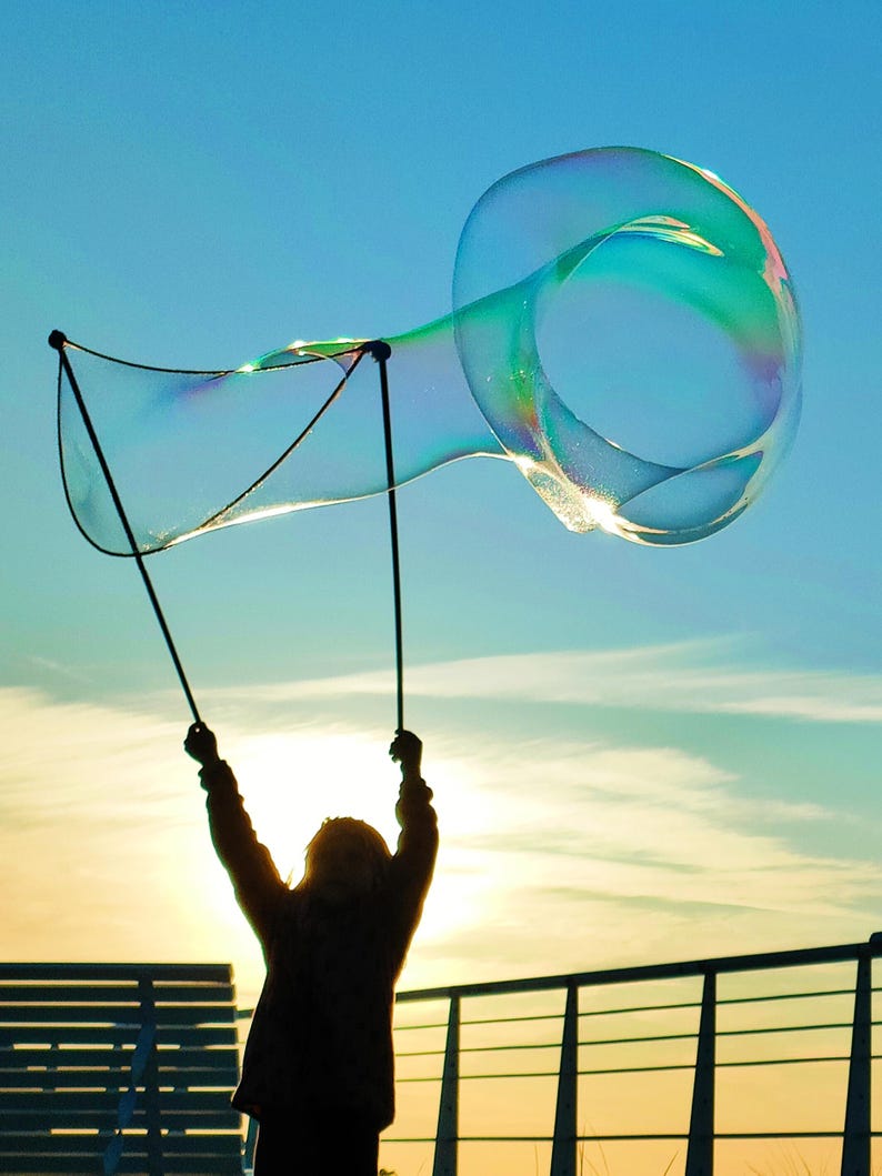 May include: A large, iridescent bubble floats in the sky, held aloft by a person using a bubble wand. The bubble is a vibrant display of colors against a blue sky, with the sun setting in the background. The scene evokes a sense of joy and play.