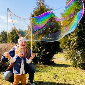 May include: A large, iridescent bubble floats in the air, held by two wooden sticks. A man and a child are holding the sticks, smiling. The bubble reflects the colors of the rainbow. The scene takes place outdoors on a sunny day.