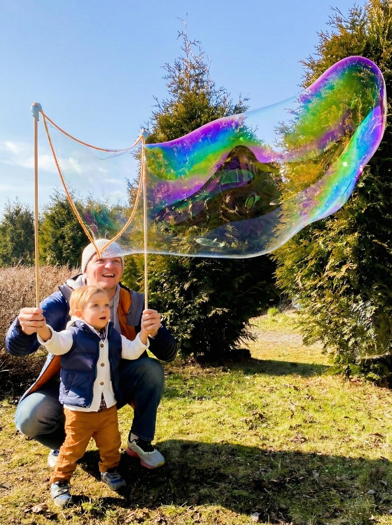 May include: A large, iridescent bubble floats in the air, held by two wooden sticks. A man and a child are holding the sticks, smiling. The bubble reflects the colors of the rainbow. The scene takes place outdoors on a sunny day.