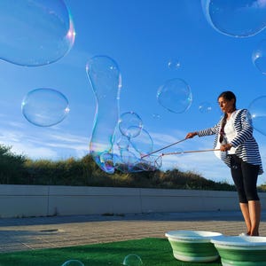 May include: A woman is making giant bubbles with a bubble wand on a sunny day. Large, transparent bubbles float in the blue sky. The woman is wearing a striped jacket and dark pants. Two green and white buckets are in the foreground.