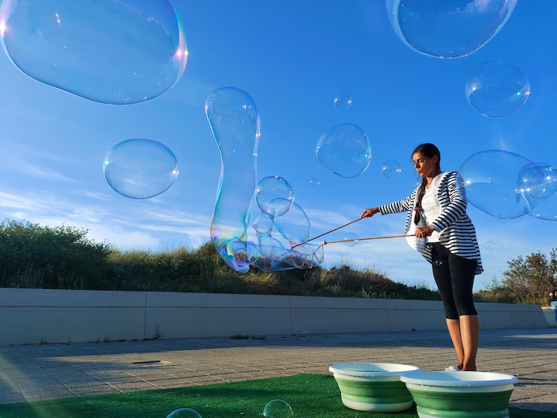 May include: A woman is making giant bubbles with a bubble wand on a sunny day. Large, transparent bubbles float in the blue sky. The woman is wearing a striped jacket and dark pants. Two green and white buckets are in the foreground.
