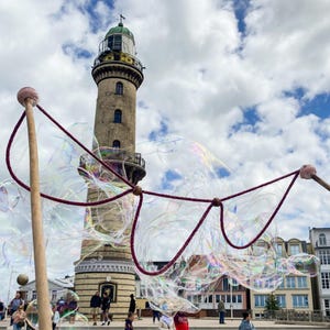 May include: A bubble wand with two wooden handles and a thick, dark red cord forming a large loop, creating iridescent soap bubbles in the foreground. In the background, a tall, cylindrical stone lighthouse stands under a cloudy blue sky.