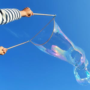 Può includere: Una grande bolla di sapone iridescente si forma da una bacchetta di legno con due manici e un cappio di corda. La bolla mostra colori arcobaleno vorticosi contro un cielo azzurro limpido. La bacchetta è tenuta da braccia con una maglia a righe.