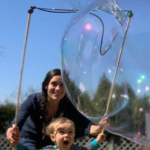 May include: A large, iridescent bubble is held aloft by two wooden wands. A woman and a child are in the background, both smiling. The child is wearing a striped shirt and appears excited. The sky is a clear blue.
