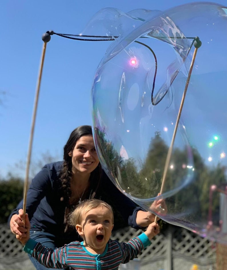 May include: A large, iridescent bubble is held aloft by two wooden wands. A woman and a child are in the background, both smiling. The child is wearing a striped shirt and appears excited. The sky is a clear blue.
