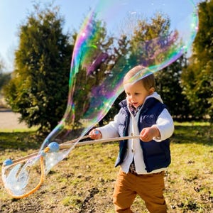 May include: A young child in a blue vest and brown pants plays with a large, iridescent bubble wand outdoors. The bubble is a large, colorful sphere. The child is holding the wand with both hands.