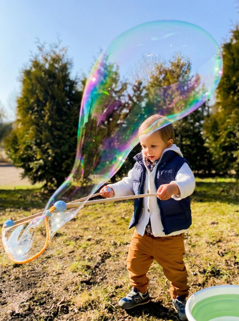 May include: A young child in a blue vest and brown pants plays with a large, iridescent bubble wand outdoors. The bubble is a large, colorful sphere. The child is holding the wand with both hands.