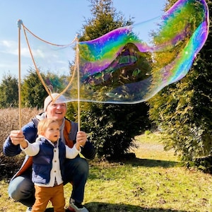 May include: A giant bubble wand consisting of two long wooden sticks connected by a flexible orange cord, creating a large, iridescent, soap bubble film with swirling rainbow colours that shimmer in the sunlight against a natural outdoor setting.
