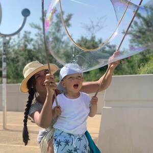 May include: A woman and a child are holding a large bubble wand, creating a giant bubble. The child is wearing a white t-shirt and blue shorts with a palm tree pattern. The woman is wearing a straw hat and a dress.