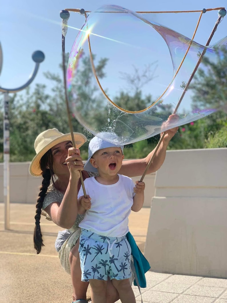 May include: A woman and a child are holding a large bubble wand, creating a giant bubble. The child is wearing a white t-shirt and blue shorts with a palm tree pattern. The woman is wearing a straw hat and a dress.