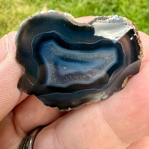 May include: A polished agate slice featuring concentric bands of deep black, translucent gray, and white. The stone has a smooth, glossy surface with a natural, irregular outer edge and a crystalline center, showcasing distinct mineral layers.