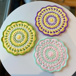 May include: Three handmade crochet coasters in shades of yellow, green, purple, and pink. Each coaster features a circular design with a scalloped edge. The coasters are placed on a white surface, showcasing the vibrant colors and intricate patterns.