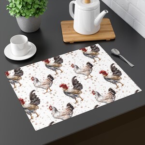 May include: A rectangular placemat featuring a repeating pattern of watercolor chickens with red combs and black tail feathers on a white background with brown speckles. The placemat is on a dark surface.