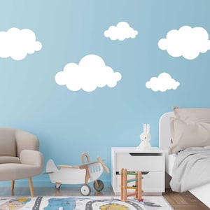 Pode incluir: Uma parede azul claro com adesivos de nuvens brancas. Uma poltrona bege fica em frente à parede. Um avião de brinquedo de madeira branco fica no chão em frente à poltrona. Uma mesa de cabeceira branca com uma escada de brinquedo de madeira fica ao lado de uma cama branca com uma cabeceira bege e um cobertor bege.