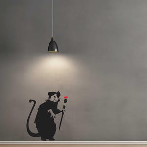 Puede incluir: Silueta negra de una rata sosteniendo un pincel con punta roja, contra una pared gris. Una lámpara colgante negra cuelga del techo, iluminando la rata. La obra de arte es de estilo plantilla.