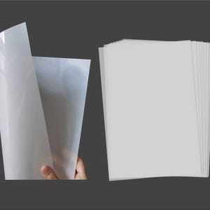 Puede incluir: Una pila de hojas de papel blancas y translúcidas. Las hojas están dobladas por la mitad y están colocadas planas sobre una superficie gris.