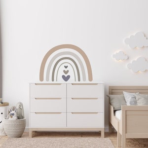 Könnte beinhalten: Eine weiße Kommode mit sechs Schubladen steht in einem Kinderzimmer mit einem hellbraunen Kleinkindbett, einem braunen Vorhang und einer Wand, die mit einem beige-braunen Regenbogen mit lila Herzen und weißer Wolkenwanddekoration verziert ist.