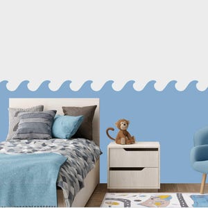 Puede incluir: Un diseño de olas azul y blanco en una pared sobre una cama individual con un edredón estampado en azul y gris. Una pequeña mesita de noche de madera con dos cajones está al lado de la cama. Un mono de peluche está sentado encima de la mesita de noche. Un sillón azul está en el fondo.