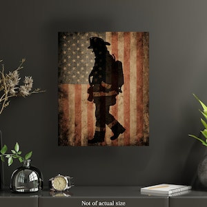 May include: A framed print featuring a silhouette of a firefighter walking in front of a distressed American flag. The artwork is predominantly black, red, and white, with a vintage, worn appearance. The text at the bottom reads "Not of actual size."