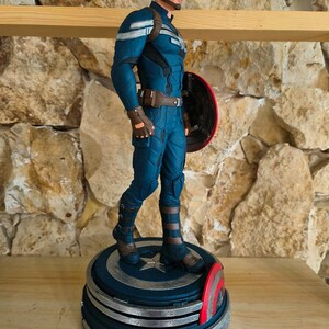 May include: A Captain America figurine in a dynamic pose, clad in a blue and silver suit, stands on a detailed base. The figure holds a red, white, and blue shield. The base is a layered design with star accents, showcasing the iconic superhero.