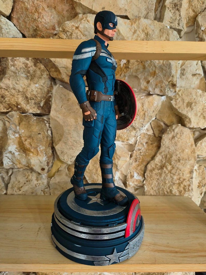 May include: A Captain America figurine in a dynamic pose, clad in a blue and silver suit, stands on a detailed base. The figure holds a red, white, and blue shield. The base is a layered design with star accents, showcasing the iconic superhero.