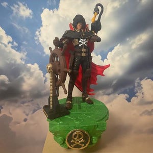 May include: A detailed figurine of a character in a black outfit with a red cape, holding a black swan and a ship's wheel. The figure stands on a green base with a gold skull and crossbones emblem, set against a cloudy sky backdrop.