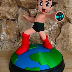 Può includere: Una statuetta del personaggio Astro Boy con capelli neri, fascia in vita verde scuro e stivali rossi, in piedi su una base a forma di globo. La figura ha un braccio teso in avanti con un dettaglio circolare di luce blu e nera sul polso.