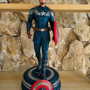 May include: A Captain America figurine in a dynamic pose, clad in a blue and silver suit, holding a red and silver shield. The figure stands on a circular base with a shield design and the words "Captain America" on it.