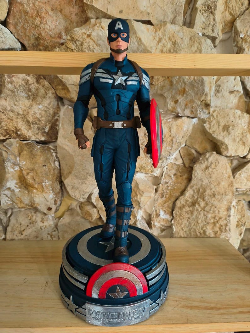 May include: A Captain America figurine in a dynamic pose, clad in a blue and silver suit, holding a red and silver shield. The figure stands on a circular base with a shield design and the words "Captain America" on it.