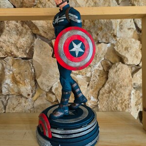 May include: A Captain America figurine in a dynamic pose, holding his iconic shield. The figure is clad in a blue and silver suit, with a matching helmet and mask. The shield is red, white, and blue, with a central star design. The figurine stands on a detailed base.