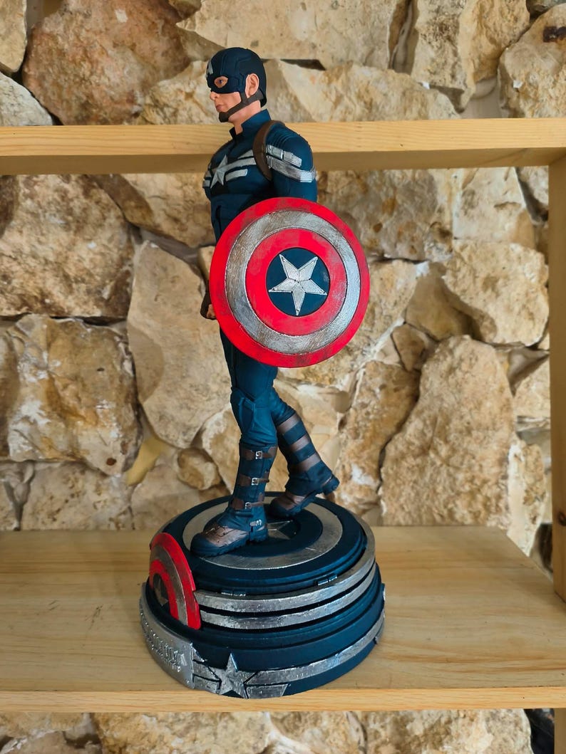 May include: A Captain America figurine in a dynamic pose, holding his iconic shield. The figure is clad in a blue and silver suit, with a matching helmet and mask. The shield is red, white, and blue, with a central star design. The figurine stands on a detailed base.