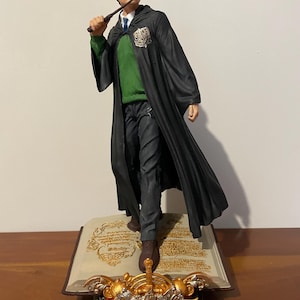 以下が含まれることがあります： 本型の台座の上に立っている黒と緑のハリーポッターのフィギュア。フィギュアは杖を持ち、眼鏡をかけています。台座には金色のホグワーツの紋章が飾られています。