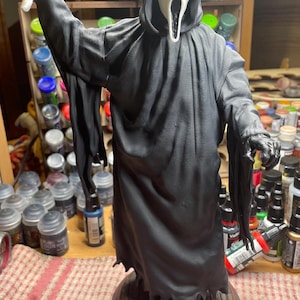 Könnte beinhalten: Eine Ghostface-Figur in einer langen schwarzen Kapuzenrobe mit einer weißen Maske, die einen weit geöffneten Mund und Augenhöhlen aufweist. Die Figur hält ein silberfarbenes Messer in der erhobenen rechten Hand und steht auf einem runden Holzsockel.