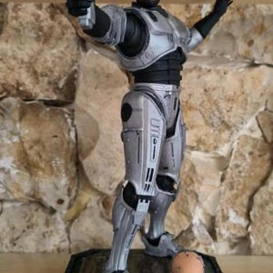 Może przedstawiać: Srebrno-czarna figurka robota stojąca na prostokątnej podstawie, trzymająca w prawej dłoni srebrny pistolet. Oddzielna głowa o ludzkich rysach spoczywa na podstawie obok stóp figurki, które są osłonięte metalowym pancerzem.
