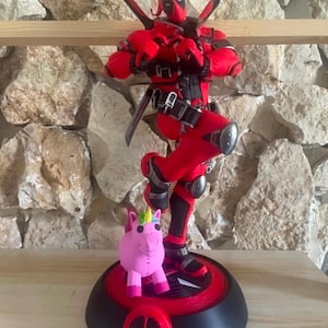 Op de afbeelding: Een Deadpool-actiefiguur in een rood en zwart pak met twee zwaarden op de rug, die met de handen een hartvorm maakt. Het figuur staat op een ronde zwarte voet met een Deadpool-logo, naast een klein roze eenhoornspeeltje met een regenbooghoorn.