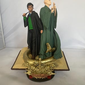 May include: A Harry Potter figurine set featuring Harry Potter and Lord Voldemort. Harry is holding his wand and wearing his Hogwarts uniform. Voldemort is wearing a green robe and is holding his wand. The figurines are standing on a golden book with the Harry Potter logo.