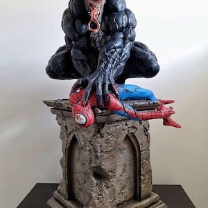 Op de afbeelding: Een gedetailleerd harsbeeld van het donkere, gespierde personage Venom dat over een verslagen Spider-Man-figuur hurkt. Beide figuren staan op een verweerde stenen sokkel met boogvormige architecturale details en barsten in het oppervlak.