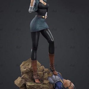 Könnte beinhalten: Eine detaillierte Sammlerfigur zeigt eine blonde Figur in schwarzem Oberteil, Jeansweste und Rock, die auf einem felsigen Sockel über einer besiegten Figur in blau-weißer Rüstung steht. Die Figur trägt eine schwarze Leggings und braune Stiefel.