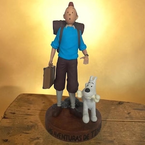 Könnte beinhalten: Eine Figur von Tim, gekleidet in einem blauen Hemd und braunen Hosen, mit einem Koffer und Rucksack. Struppi, der weiße Hund, steht neben ihm und hält einen Knochen. Der Sockel ist braun mit Text am Rand.