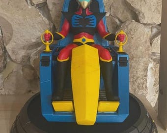 Puede incluir: Figura detallada de un personaje de la serie de anime Uforobot Grendizer, sentado en una silla azul. La figura es roja, negra y amarilla, con un casco dorado. La base es gris con el logo del programa.